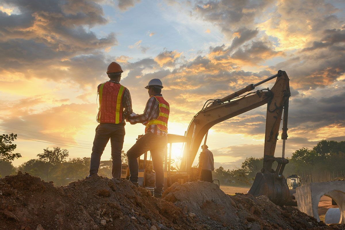 Construction site, two engineers review plans with an excavator in the background.